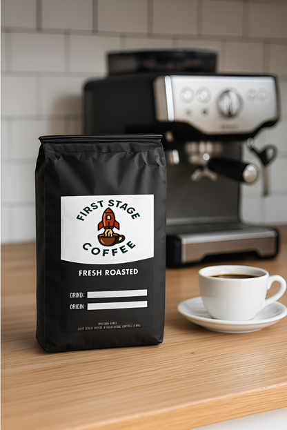 Black coffee bag with 'First Stage Coffee' branding on a kitchen counter with a coffee machine and cup.